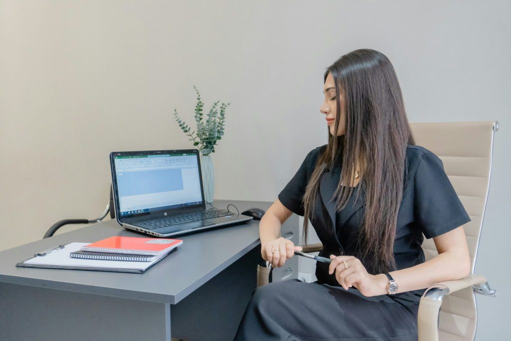 Woman in black dress working at desk with laptop.